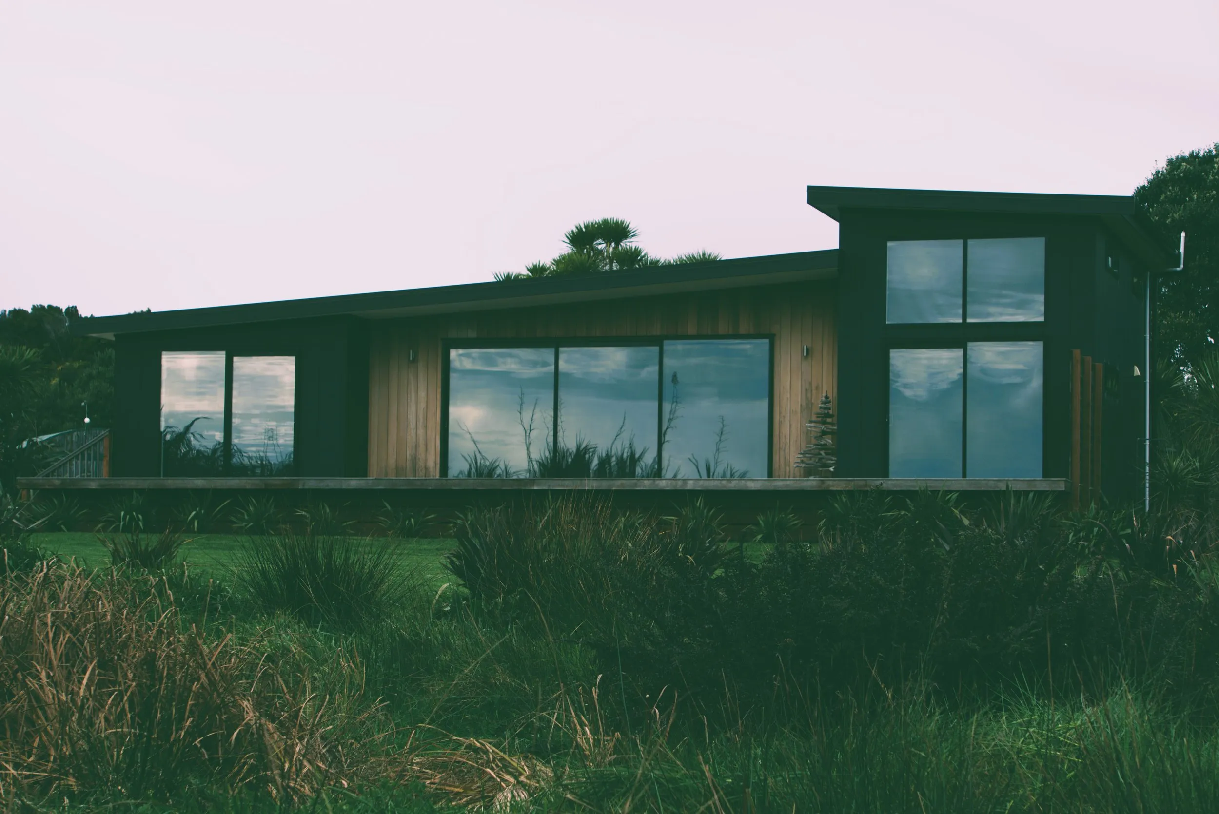 The full elevation at dusk — mono-pitch roof, dark cladding with a cedar centre bay, and full-height glazing onto the deck