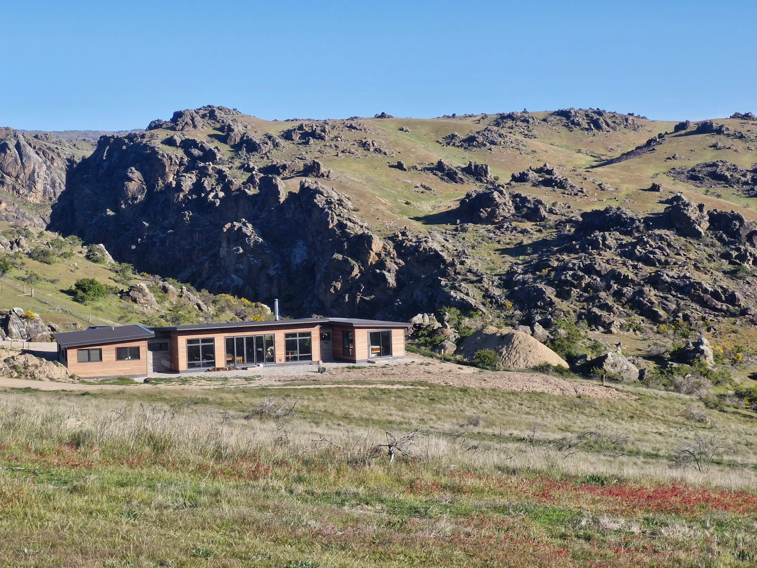 The house sitting low in tussock and schist country, with the rocky hillside rising behind it